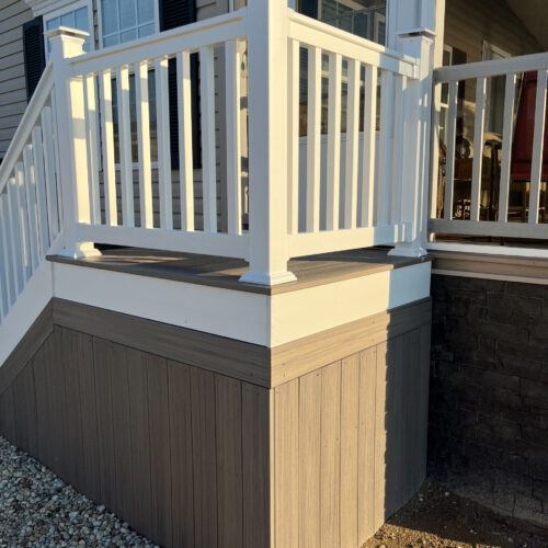 Close-up of a custom-built deck corner featuring gray composite decking, white railing posts, and a stone veneer foundation.