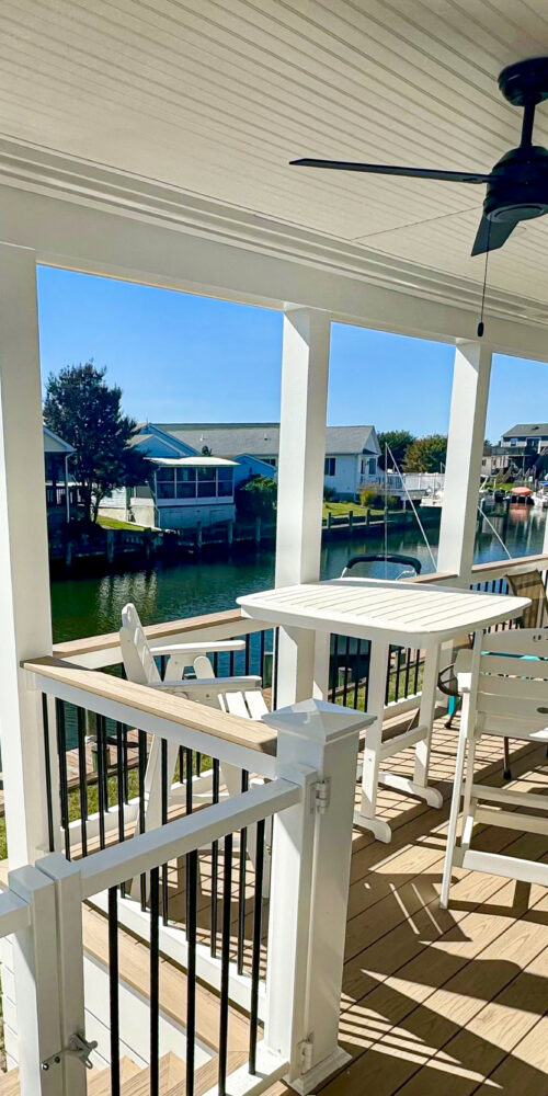 A luxury covered waterfront porch featuring a natural wood tongue-and-groove ceiling, black ceiling fans, and white railings overlooking a dock.
