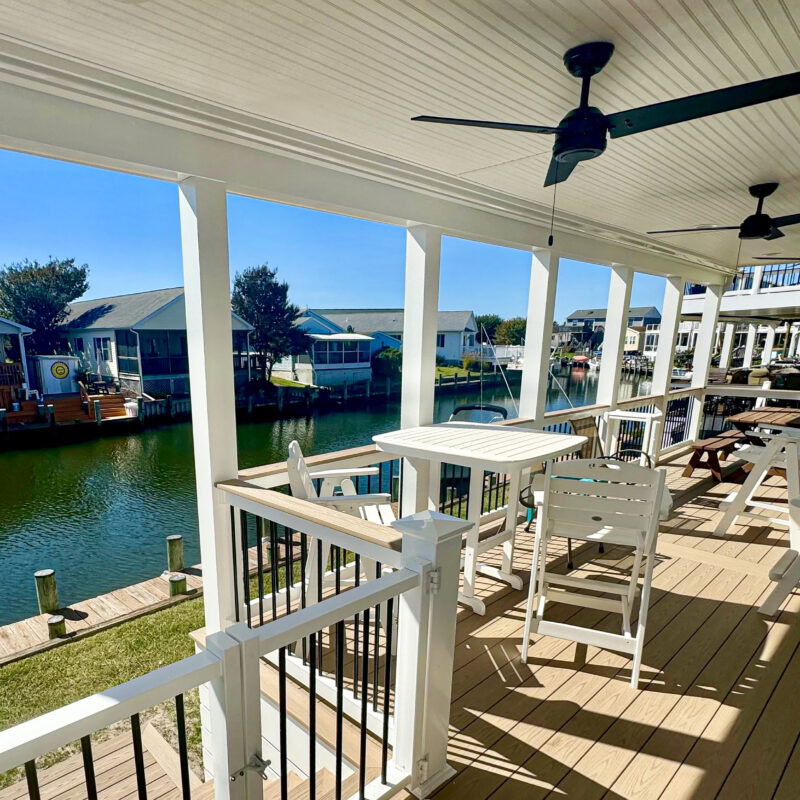 A luxury covered waterfront porch featuring a natural wood tongue-and-groove ceiling, black ceiling fans, and white railings overlooking a dock.