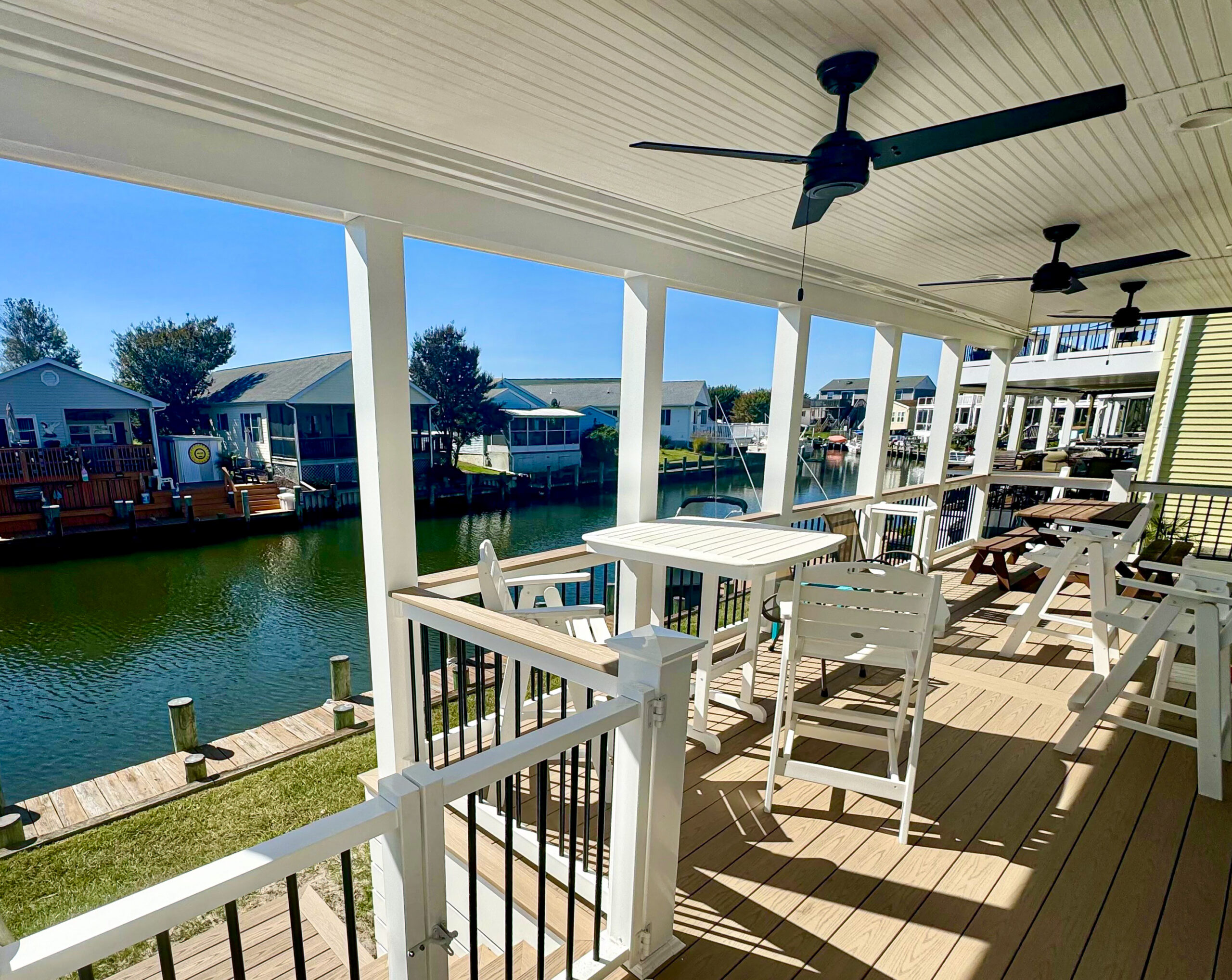 A luxury covered waterfront porch featuring a natural wood tongue-and-groove ceiling, black ceiling fans, and white railings overlooking a dock.