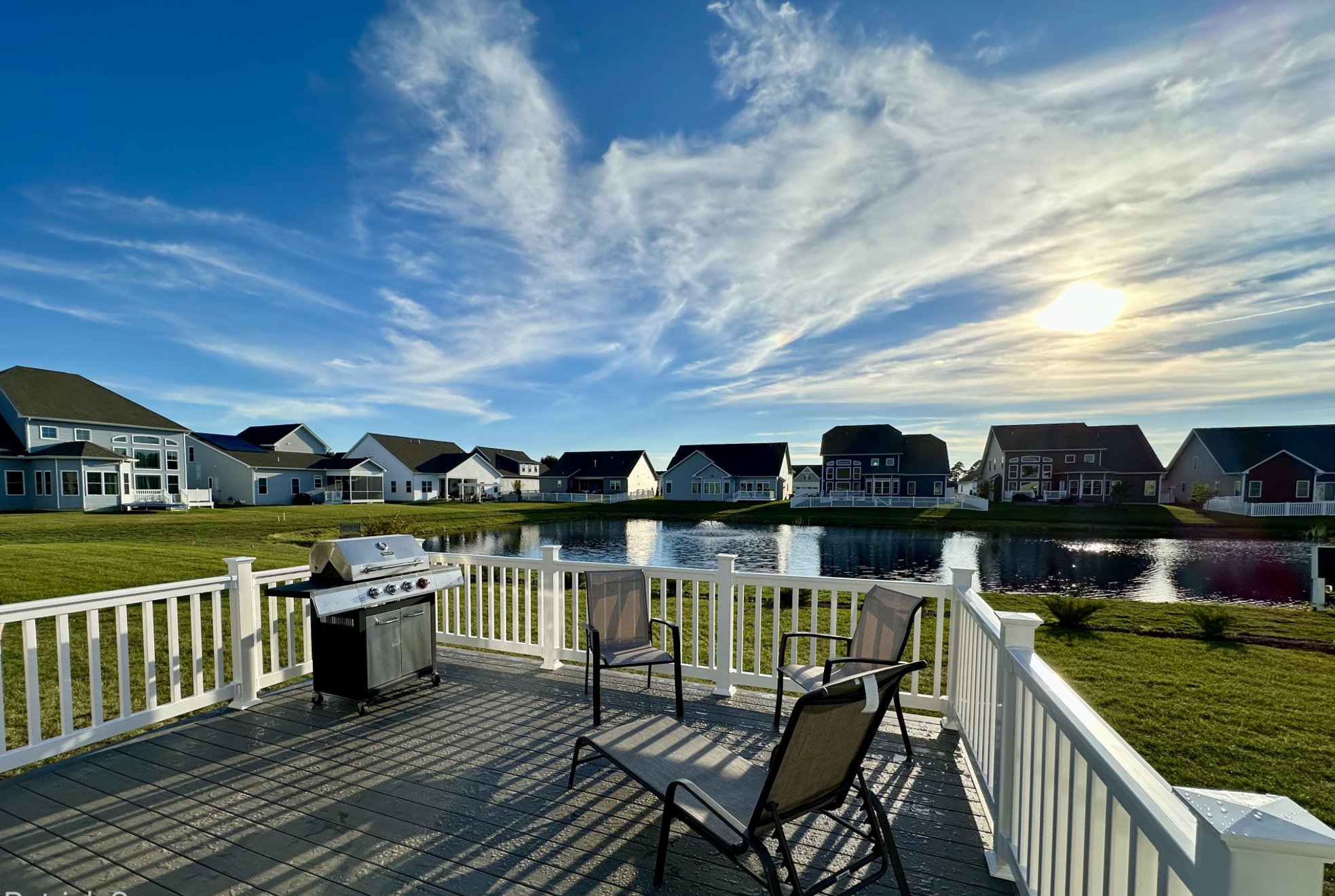 Sunset POV of a grey TimberTech composite deck with white railings and a grill, overlooking a neighborhood pond.