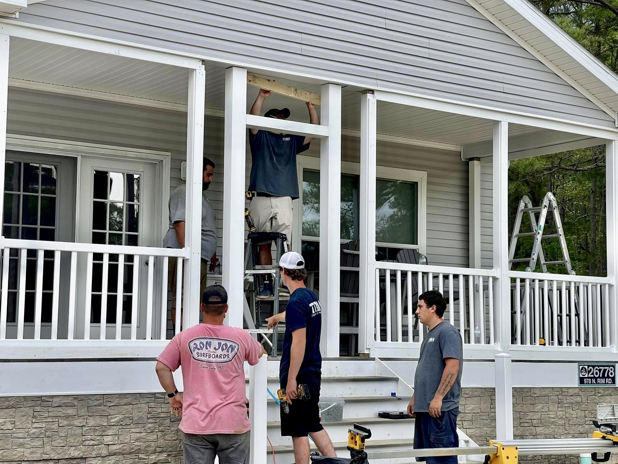 Kyle Speese, owner of Titan Decks, wearing a dark blue shirt and pointing while directing a crew member on a ladder during a porch build.
